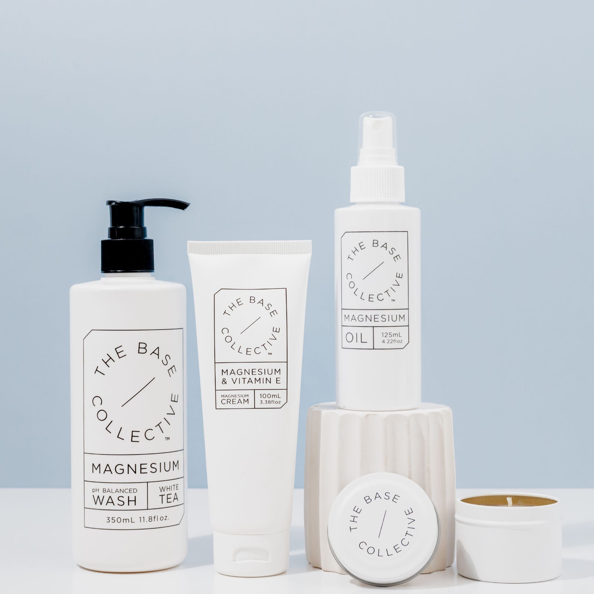 Magnesium Body Wash, Magnesium Cream with Vitamin E, Magnesium Oil Spray on white pillar with scented soy candle open next to it against light sky blue background.