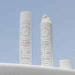 SPF50 Mineral tinted face sunscreen two bottles, with water drops on them against a blue background.